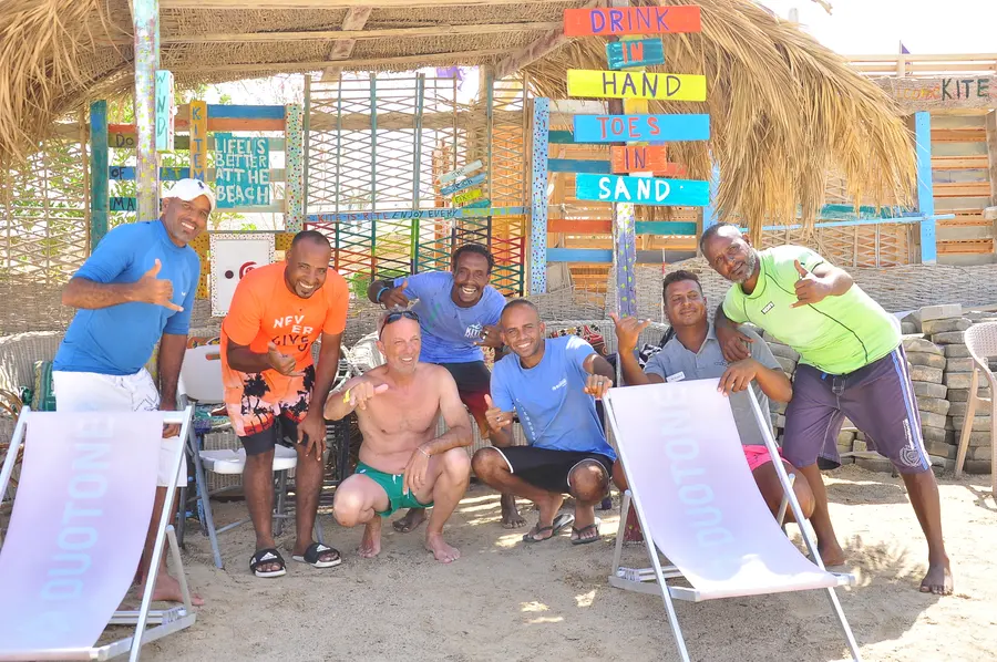 KiteMood team and guests posing together under a thatched beach shelter, several making a shaka hand sign