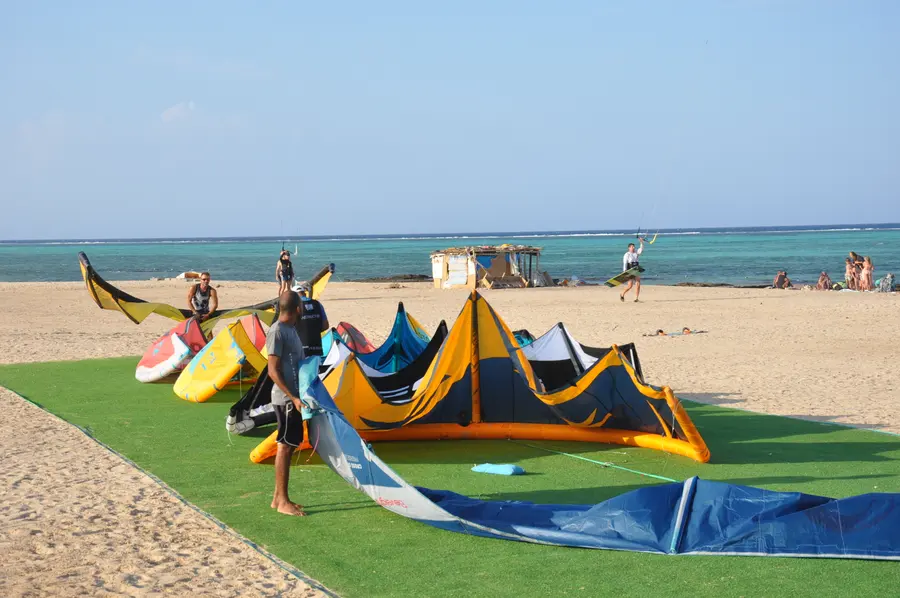 Colourful kites laid out on a green mat on the beach as people prepare equipment, with the turquoise lagoon behind