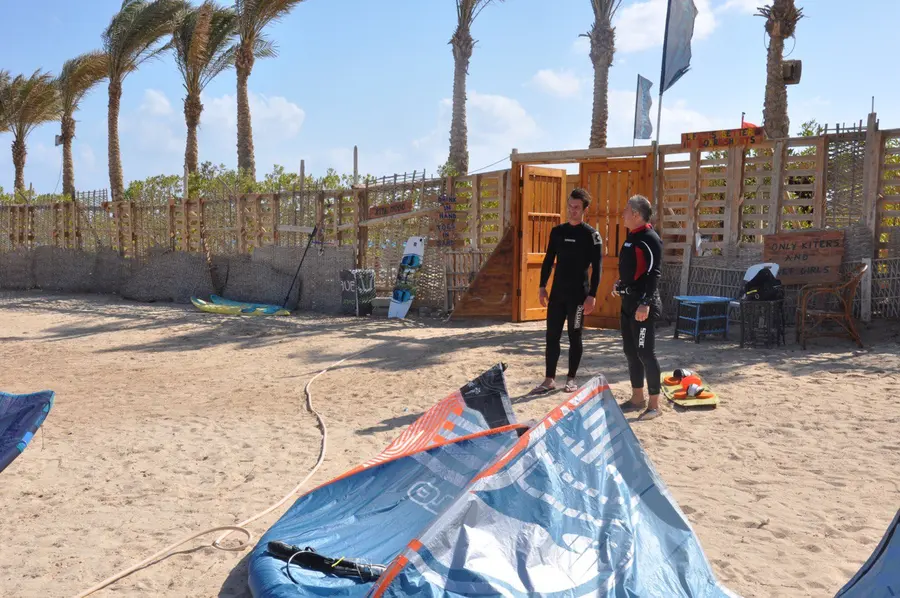 Two people in wetsuits chatting next to a large blue kite on the sand, with the wooden KiteMood base and palm trees behind