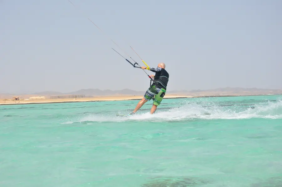 Kitesurfer seen from behind riding across the shallow turquoise lagoon, spray at the board, sandy shore and hills beyond